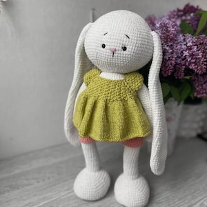 May include: A white crocheted bunny wearing a green dress and pink shorts. The bunny has long ears and a sweet expression.