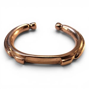 May include: A copper-colored cuff bracelet with an open design. The bracelet has a rounded shape with decorative ridges and a rectangular section in the center. The ends of the bracelet have small, rounded details.