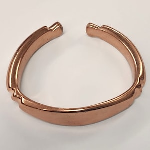 May include: A copper-colored cuff bracelet with an open design. The bracelet has a smooth, curved band with decorative notches on the sides. The ends of the bracelet are slightly squared off.
