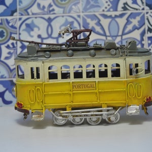 May include: A yellow and white metal model tram featuring the word PORTUGAL printed on the side. The vehicle has a grey roof with a pantograph, multiple windows, and silver-colored wheels with visible mechanical linkages along the lower chassis.