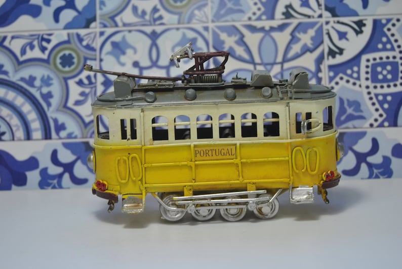 May include: A yellow and white metal model tram featuring the word PORTUGAL printed on the side. The vehicle has a grey roof with a pantograph, multiple windows, and silver-colored wheels with visible mechanical linkages along the lower chassis.