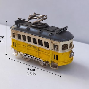 May include: A yellow and white model tram with a dark gray roof, featuring the text "PORTUGAL" and "GRA&Ccedil;A 28". The tram measures 2.0 inches in height and 3.5 inches in length, including a metal pantograph on top and small wheels at the base.