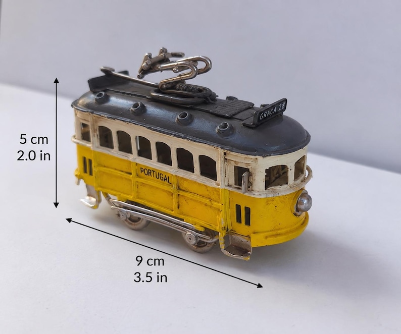 May include: A yellow and white model tram with a dark gray roof, featuring the text "PORTUGAL" and "GRA&Ccedil;A 28". The tram measures 2.0 inches in height and 3.5 inches in length, including a metal pantograph on top and small wheels at the base.