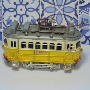 May include: A yellow and white metal model tram featuring the word LISBOA on the side. The vehicle has a grey roof, a brown pantograph on top, multiple windows, and small silver wheels. The design includes red taillights and silver headlights.