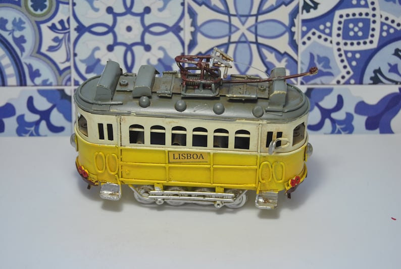 May include: A yellow and white metal model tram featuring the word LISBOA on the side. The vehicle has a grey roof, a brown pantograph on top, multiple windows, and small silver wheels. The design includes red taillights and silver headlights.