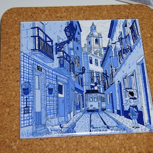 May include: A square ceramic coaster with a blue and white illustration of a European street scene, featuring a tram on tracks. The coaster is set on a brown cork base.