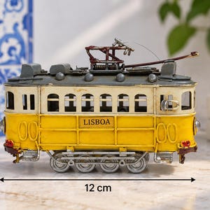 May include: A yellow and cream-colored model tram with the word LISBOA printed on the side. The tram features a dark gray roof, a metal pantograph, and four silver wheels. It measures 7 cm in height and 12 cm in length as indicated by arrows.