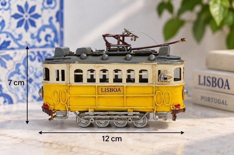 May include: A yellow and cream-colored model tram with the word LISBOA printed on the side. The tram features a dark gray roof, a metal pantograph, and four silver wheels. It measures 7 cm in height and 12 cm in length as indicated by arrows.