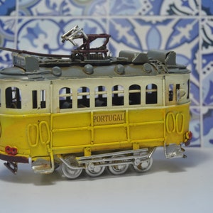 May include: A yellow and white metal model tram featuring the word PORTUGAL printed on the side. The toy vehicle has a grey roof, a dark brown pantograph on top, silver-colored wheels, and a front headlight, capturing the look of a classic streetcar.