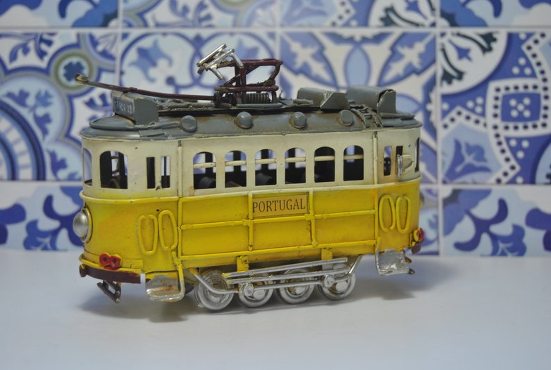 May include: A yellow and white metal model tram featuring the word PORTUGAL printed on the side. The toy vehicle has a grey roof, a dark brown pantograph on top, silver-colored wheels, and a front headlight, capturing the look of a classic streetcar.