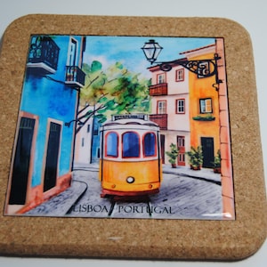 May include: A cork coaster featuring a vibrant illustration of a yellow tram on a cobbled street in Lisbon, Portugal. The buildings are painted in blue and orange. The text "LISBOA PORTUGAL" is visible at the bottom of the image.