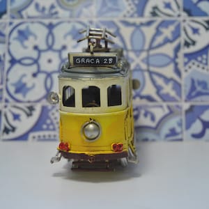 May include: A yellow and white metal model tram featuring a front sign that reads GRACA 28. The miniature vehicle includes a silver headlight, red taillights, a dark brown bumper, and a metal pantograph on the roof, capturing the classic design.