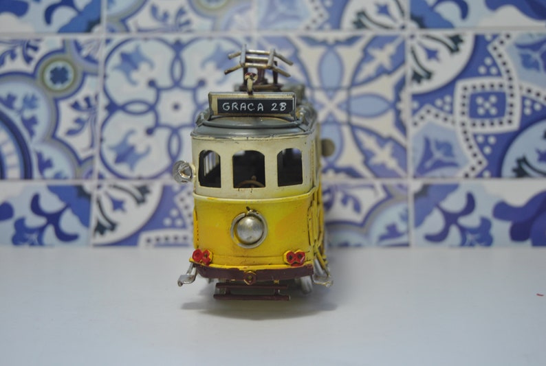 May include: A yellow and white metal model tram featuring a front sign that reads GRACA 28. The miniature vehicle includes a silver headlight, red taillights, a dark brown bumper, and a metal pantograph on the roof, capturing the classic design.