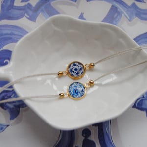 Può includere: Due delicati braccialetti con ciondoli rotondi a motivi blu e bianchi incastonati in lunette color oro. I braccialetti poggiano su un piatto bianco a forma di foglia, su uno sfondo di motivi floreali blu e bianchi. I braccialetti sono collegati da un sottile cordino chiaro.
