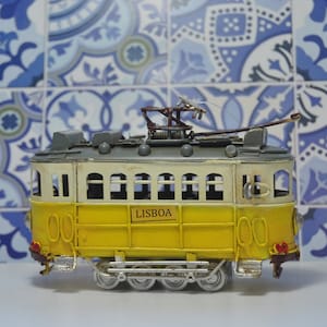 May include: A yellow and white model tram featuring the word LISBOA on its side. The vehicle has a grey roof with a metal pantograph on top, silver-colored wheels, and small red detailing on the front and rear bumpers of the carriage.