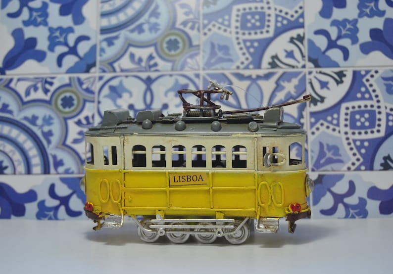 May include: A yellow and white model tram featuring the word LISBOA on its side. The vehicle has a grey roof with a metal pantograph on top, silver-colored wheels, and small red detailing on the front and rear bumpers of the carriage.