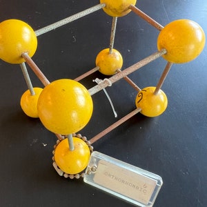 May include: A yellow model of an orthorhombic crystal structure with a tag that reads "ORTHORHOMBIC".