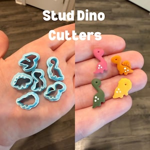 May include: A hand holds blue plastic dinosaur-shaped cookie cutters and several small, colorful dinosaur-shaped studs. The studs are pink, orange, green, and yellow. Text in the image reads "Stud Dino Cutters."