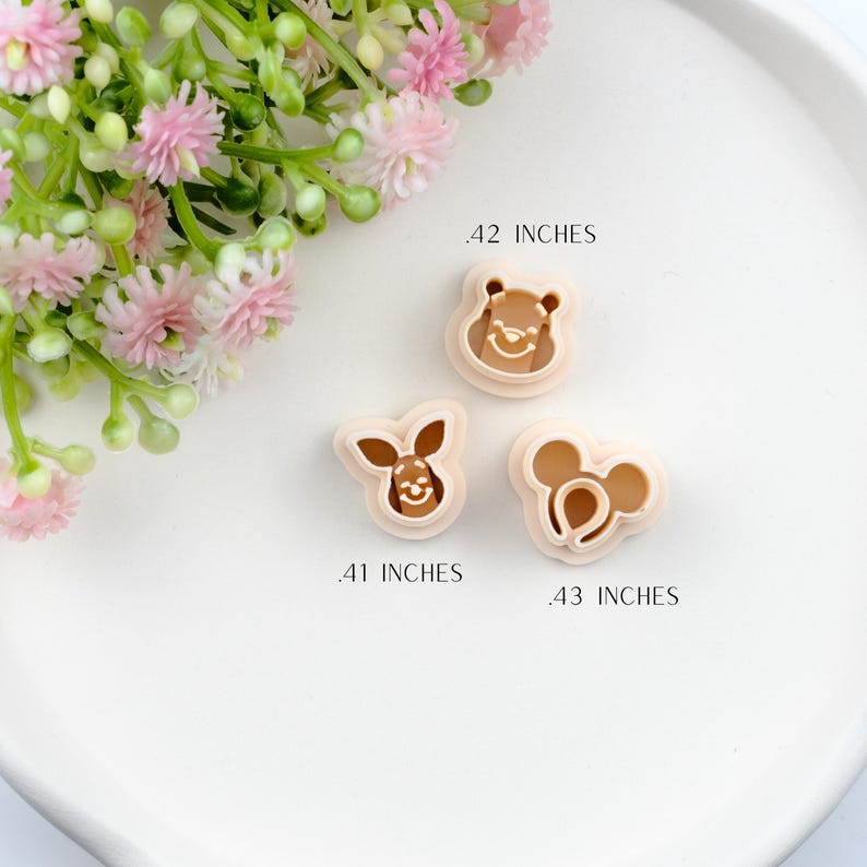 May include: Three tan-colored cookie cutters in the shapes of a bear, a rabbit, and a mouse. The bear cutter is .42 inches, the rabbit is .41 inches, and the mouse is .43 inches. The cutters are on a white surface with pink flowers.