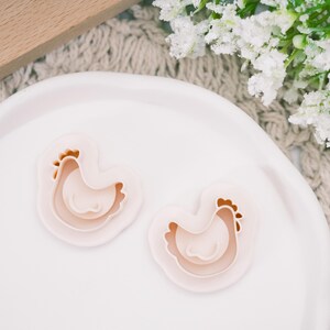 May include: Two light pink chicken-shaped cookie cutters on a white plate. The cookie cutters have a layered design with brown details for the comb and beak. The plate is on a neutral-colored surface with white flowers in the background.