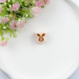 May include: A small, light brown cookie cutter in the shape of a cartoon kangaroo with a smiling face. The cutter is set against a white background with pink and green floral accents.