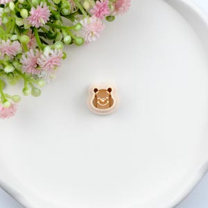 May include: A light brown bear-shaped cookie cutter with a smiling face. The cutter is made of plastic and has a detailed outline. The image is on a white surface with pink flowers in the background.