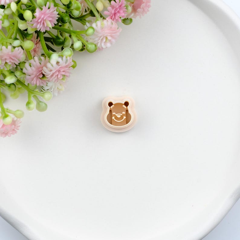 May include: A light brown bear-shaped cookie cutter with a smiling face. The cutter is made of plastic and has a detailed outline. The image is on a white surface with pink flowers in the background.