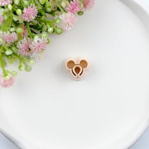 May include: A light beige plastic cookie cutter in the shape of a cartoon mouse head. The cutter has two circular ears and a rounded head shape. The cutter is on a white surface with pink flowers.