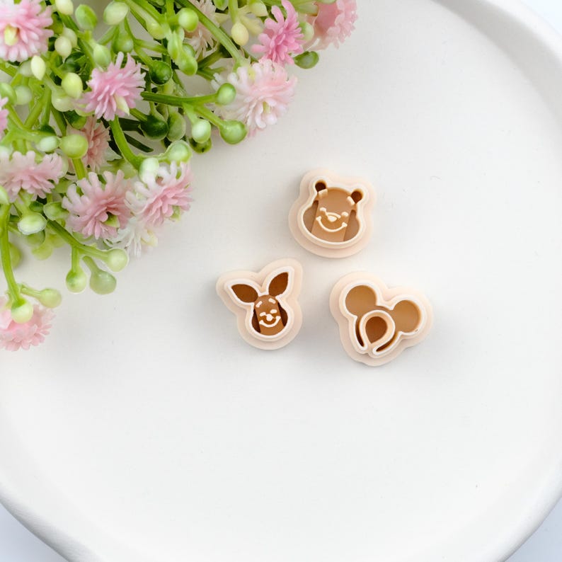 May include: Three tan-colored cookie cutters featuring cartoon character designs. The cutters include Winnie the Pooh, a rabbit, and Mickey Mouse. The cutters are on a white surface with pink flowers in the background.