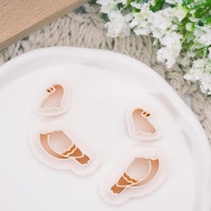 May include: A set of four light pink plastic cookie cutters in the shape of a bird and a hat. The bird cutters have detailed wing patterns. The cutters are arranged on a white plate, with white flowers in the background.
