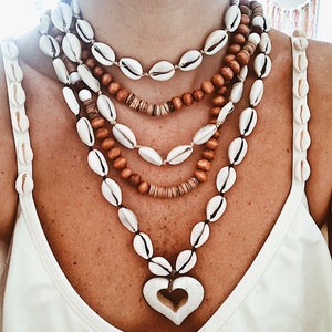 Puede incluir: Un collar de varias hebras con conchas blancas, cuentas de madera y un colgante en forma de corazón. El collar se lleva sobre una camiseta sin mangas blanca con detalles de conchas en las correas. La joyería tiene un estilo bohemio natural.