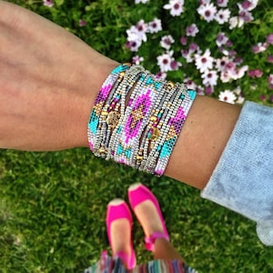 May include: A colourful beaded bracelet in silver, turquoise, pink, purple, and gold. The bracelet is worn on a wrist, with bright pink shoes visible below. The bracelet has a geometric pattern.