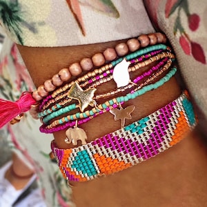 Pode incluir: Um pulso é adornado com uma coleção de pulseiras coloridas. As pulseiras apresentam vários designs, incluindo fios de contas, uma faixa tecida com um pingente de elefante e uma borla rosa. As pulseiras são empilhadas, criando um visual em camadas.