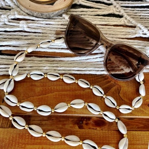 Peut inclure: Des lunettes de soleil à monture marron reposent sur une surface en bois à côté d'un collier fait de coquillages blancs et de perles dorées. Le collier est posé sur la surface en bois. Une bougie est posée sur un dessous de verre en bois à l'arrière-plan.