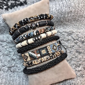 Puede incluir: Una variedad de pulseras de cuentas en varios diseños y colores, incluyendo negro, dorado, blanco y azul. Las pulseras se exhiben sobre una almohada beige contra un fondo de punto gris. Las pulseras parecen hechas a mano.