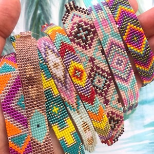 May include: A collection of beaded bracelets in various colors, including pink, orange, turquoise, and gold. Each bracelet features intricate geometric patterns. The bracelets are held in a hand against a background of palm fronds.