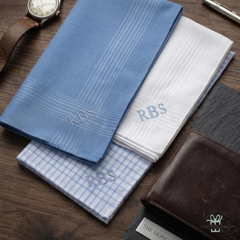 Puede incluir: Tres pa&ntilde;uelos monogramados en azul y blanco, con las iniciales "RBS" bordadas. Uno es azul claro con un patr&oacute;n de rayas, otro es blanco con un patr&oacute;n de rayas, y el tercero es azul claro con un patr&oacute;n de cuadros. Tambi&eacute;n se ve una cartera de cuero marr&oacute;n.
