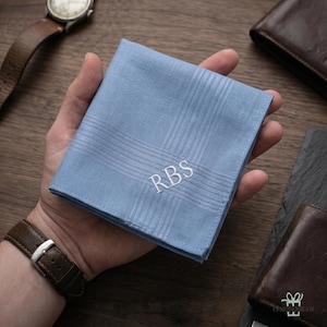 Puede incluir: Un pa&ntilde;uelo azul claro con un dise&ntilde;o a rayas y las iniciales "RBS" bordadas en blanco. El pa&ntilde;uelo se sostiene en una mano, con un reloj y una cartera de cuero visibles en el fondo.