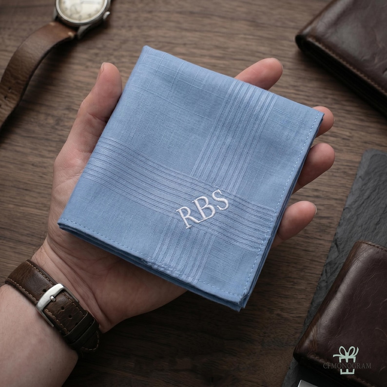 Puede incluir: Un pa&ntilde;uelo azul claro con un dise&ntilde;o a rayas y las iniciales "RBS" bordadas en blanco. El pa&ntilde;uelo se sostiene en una mano, con un reloj y una cartera de cuero visibles en el fondo.