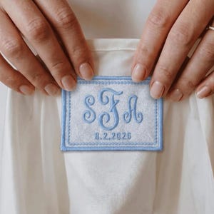 May include: A rectangular white fabric patch with a light blue embroidered border and matching light blue embroidery featuring the monogram SFA and the date 8.2.2026, stitched onto a plain white garment.