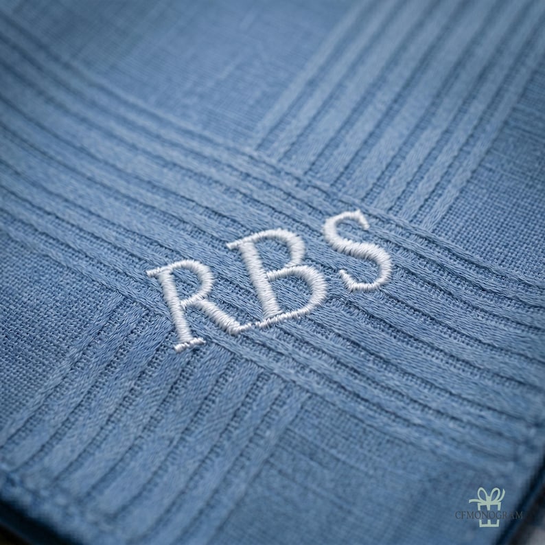 Puede incluir: Primer plano de un pa&ntilde;uelo azul claro con un dise&ntilde;o texturizado y acanalado. Las iniciales "RBS" est&aacute;n bordadas en blanco. La tela tiene un brillo sutil, lo que sugiere un material de alta calidad.