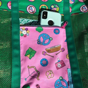 May include: A green mesh tote bag with a pink zippered pocket. The pocket has a variety of colorful Girl Scout themed designs, including peace signs, flowers, and a boat. The text "Daisy" is visible on the boat. The bag has green straps and a green mesh design. The text "Handmade by Heidi and Yvonne" is visible on the pocket.