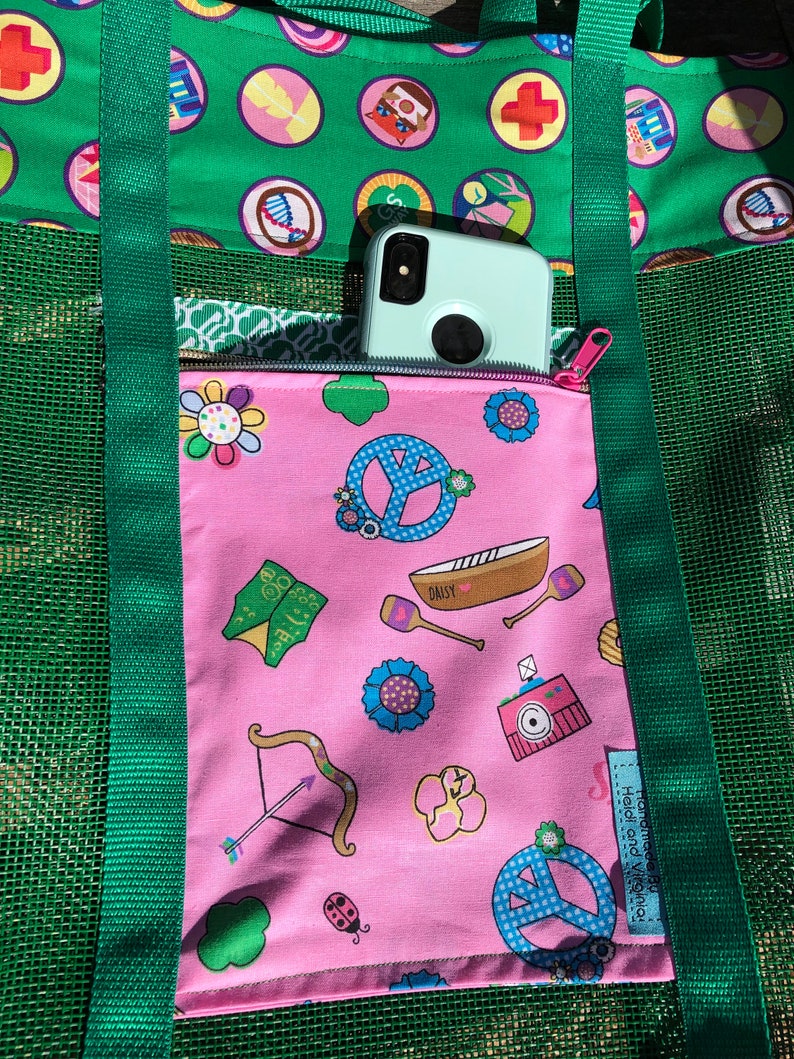 May include: A green mesh tote bag with a pink zippered pocket. The pocket has a variety of colorful Girl Scout themed designs, including peace signs, flowers, and a boat. The text "Daisy" is visible on the boat. The bag has green straps and a green mesh design. The text "Handmade by Heidi and Yvonne" is visible on the pocket.