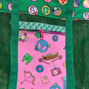 May include: A pink mesh tote bag with green straps and a zippered pocket. The pocket has a pink background with a variety of colorful images, including peace signs, flowers, and a canoe. The text "Handmade by Heidi and Virginia" is visible on the bag.