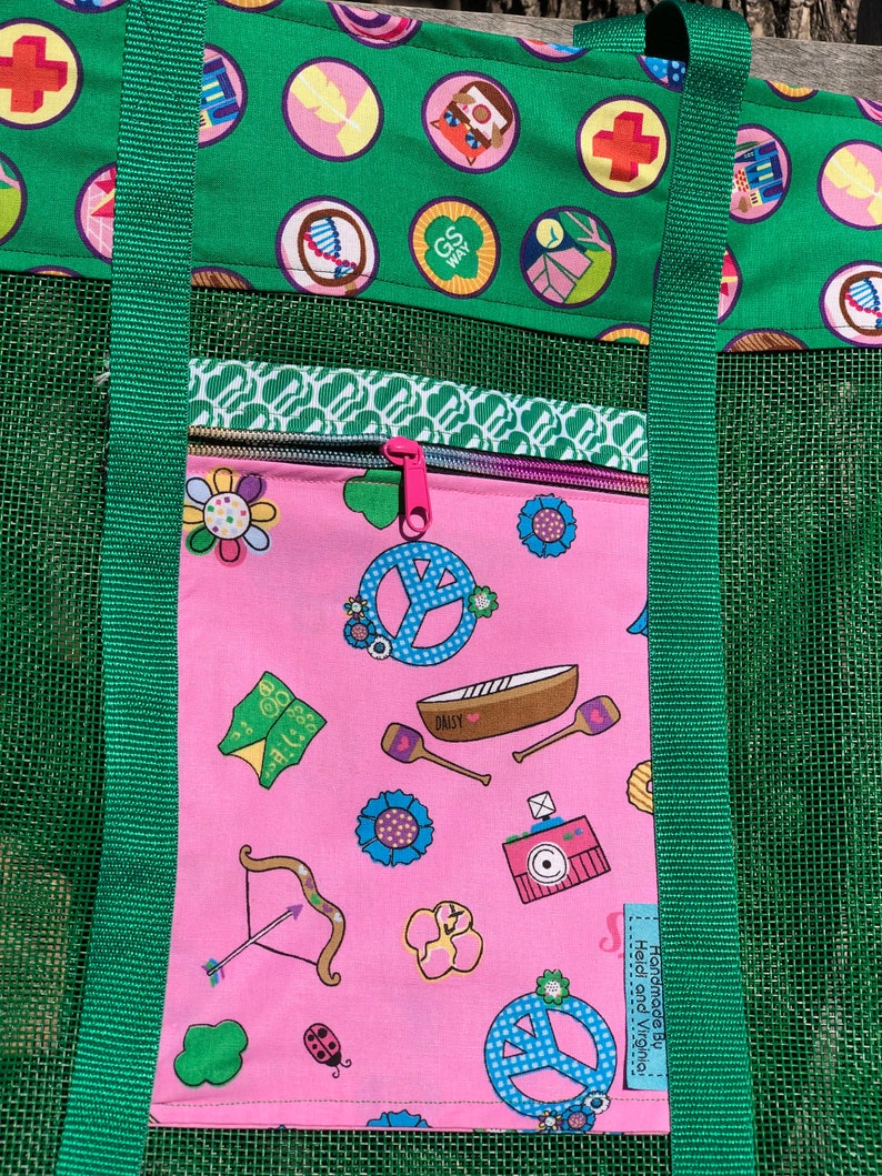May include: A pink mesh tote bag with green straps and a zippered pocket. The pocket has a pink background with a variety of colorful images, including peace signs, flowers, and a canoe. The text "Handmade by Heidi and Virginia" is visible on the bag.