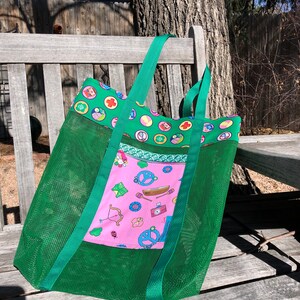May include: Green mesh tote bag with a pink fabric pocket featuring a Girl Scout theme. The pocket has a variety of images including a peace sign, a canoe, a bow and arrow, and a camera. The bag has two green straps.