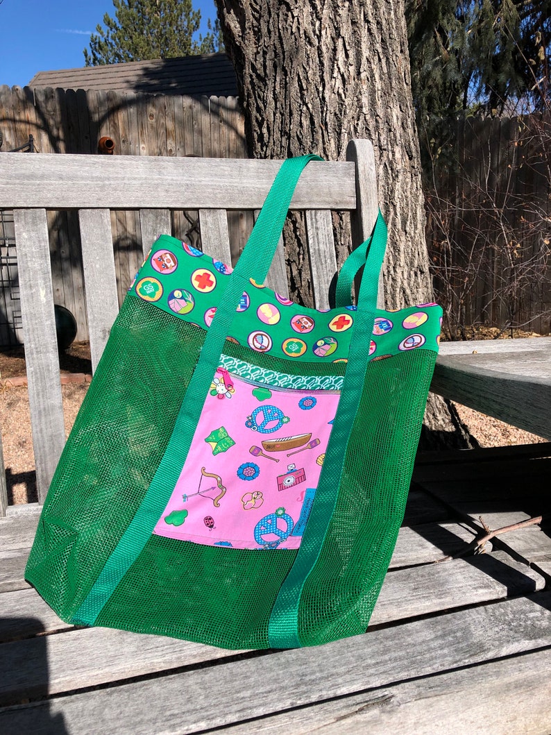 May include: Green mesh tote bag with a pink fabric pocket featuring a Girl Scout theme. The pocket has a variety of images including a peace sign, a canoe, a bow and arrow, and a camera. The bag has two green straps.