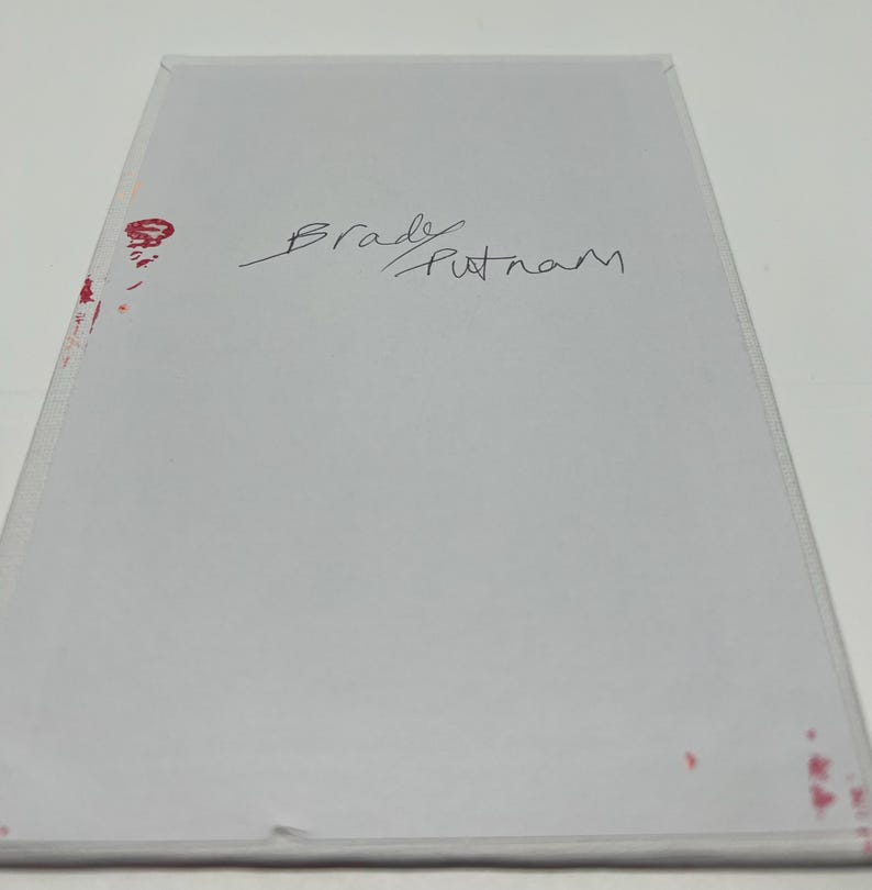 May include: White card with the handwritten signature "Brady Putnam" in black ink. There are red ink stains on the card.