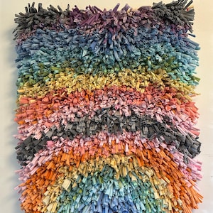 May include: A colorful, handmade wall hanging with a textured, shaggy appearance. The piece features horizontal bands of various colors, including shades of blue, green, yellow, orange, and pink. The textile art is made from fabric strips, creating a unique, tactile surface.