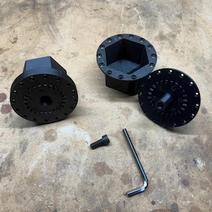 May include: Two black 3D-printed circular components with hexagonal centers and multiple perimeter holes, one additional circular plate with a central post, a single black bolt, and a silver L-shaped hex key tool arranged on a flat surface.